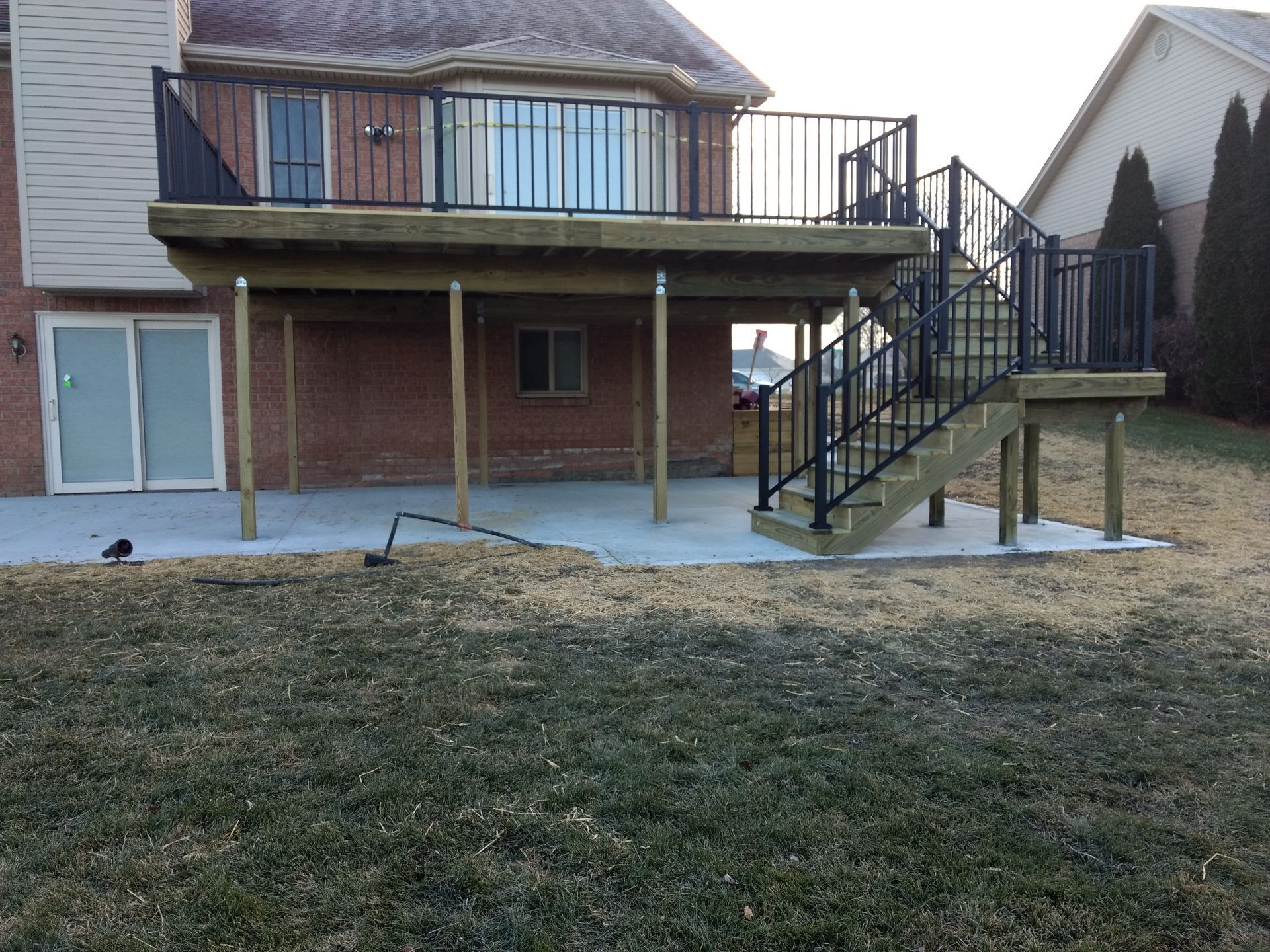 Backyard deck with stairs, black railing, supports on concrete patio, and brick house.