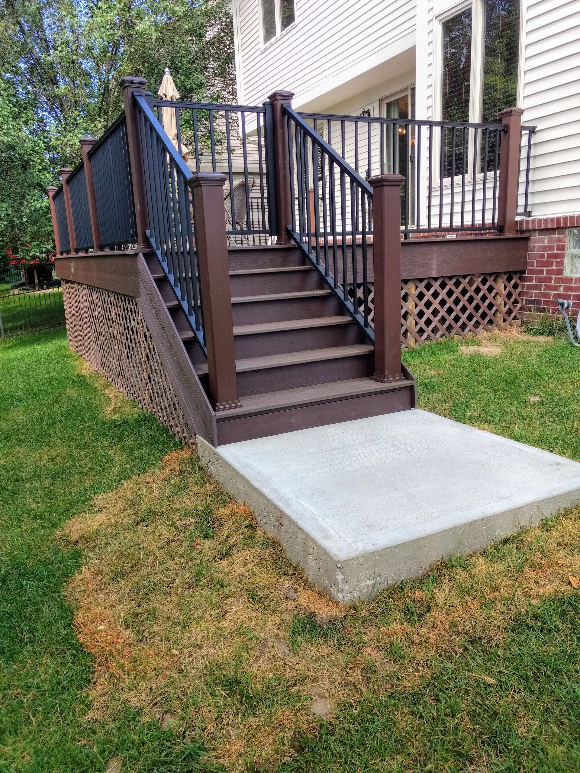 A deck with brown stairs, railing, and trim, leading to a concrete landing on a grassy lawn.