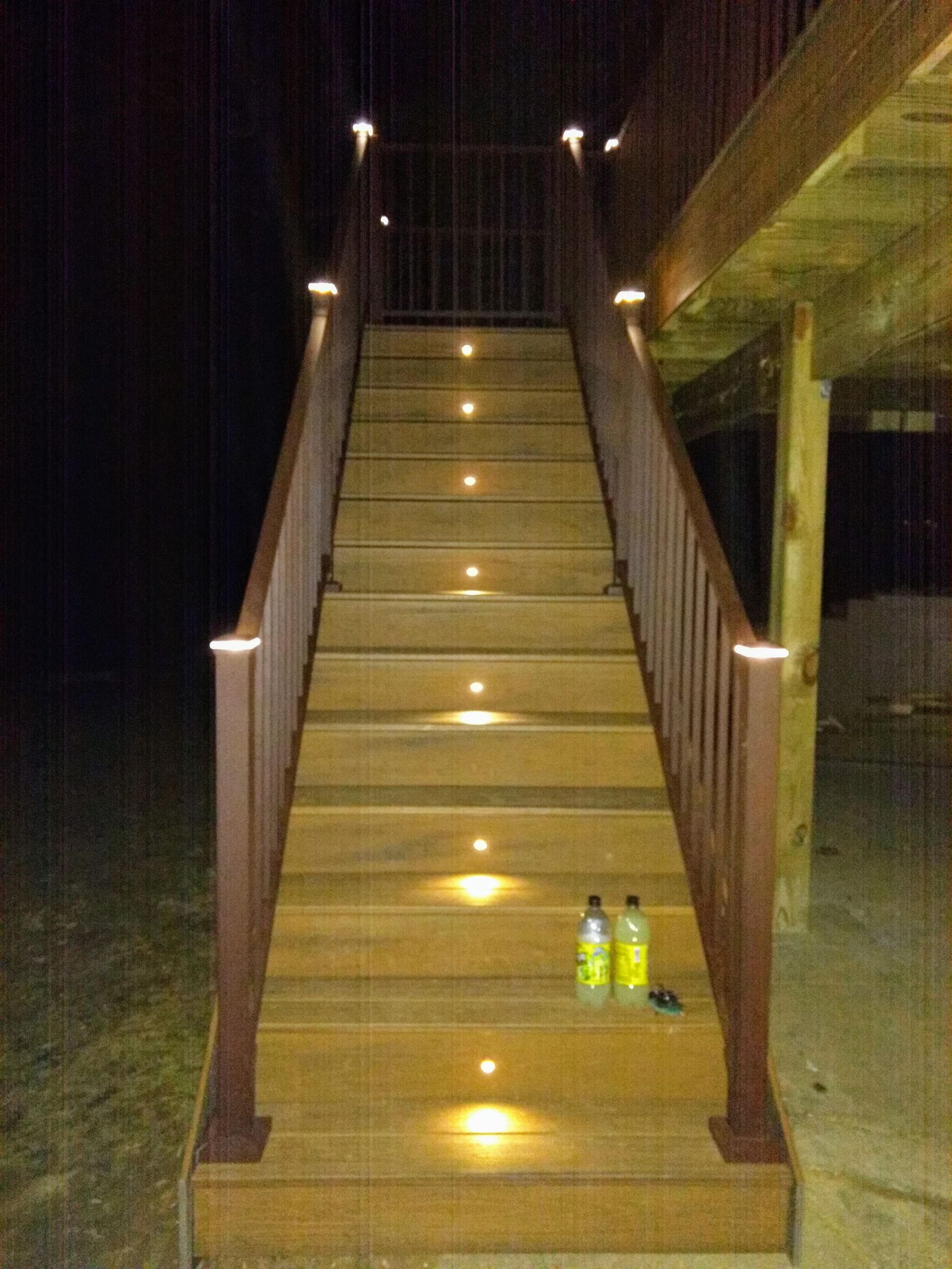 Outdoor wooden staircase at night, illuminated by recessed lights on each step and railing posts. Two bottles on the steps.