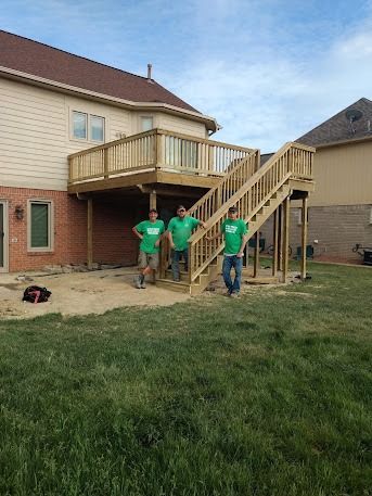 Three people stand near a newly constructed wooden deck attached to a house. They wear green shirts.