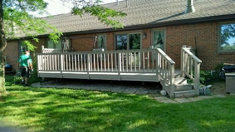 Deck with railing and steps in a backyard, beside a brick house and green grass.