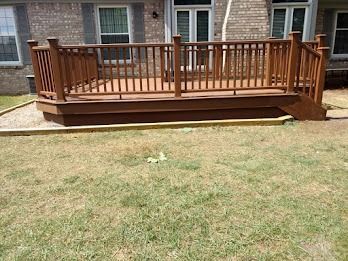 Wooden deck with railings, brown stain, attached to a brick house, sitting on a dry lawn.