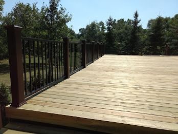 Wooden deck with brown posts and black railing, surrounded by trees under a blue sky.