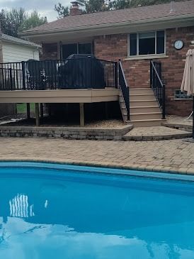 Backyard with pool, deck, and brick house. Stairs lead up to the deck with black railings.