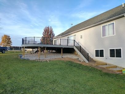 Deck attached to a two-story house with black railing, stairs, and a gravel base, set on green grass.