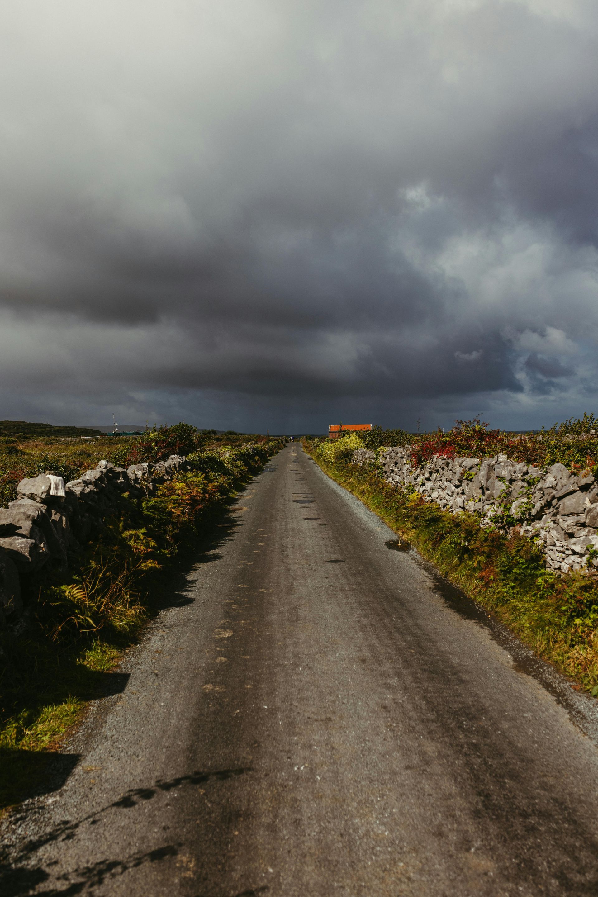 Narrow road leading towards a house under a cloudy, dark sky. Stone walls border the road.