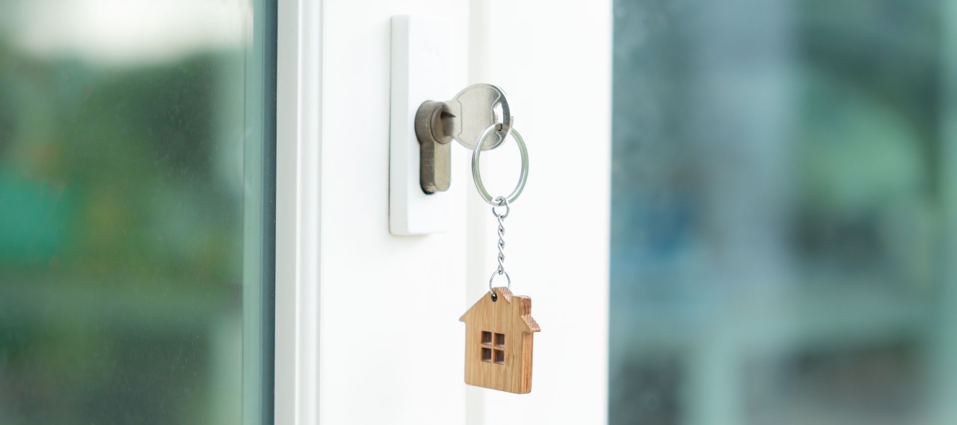 Key in a white door lock with a house-shaped keychain, blurred green background.