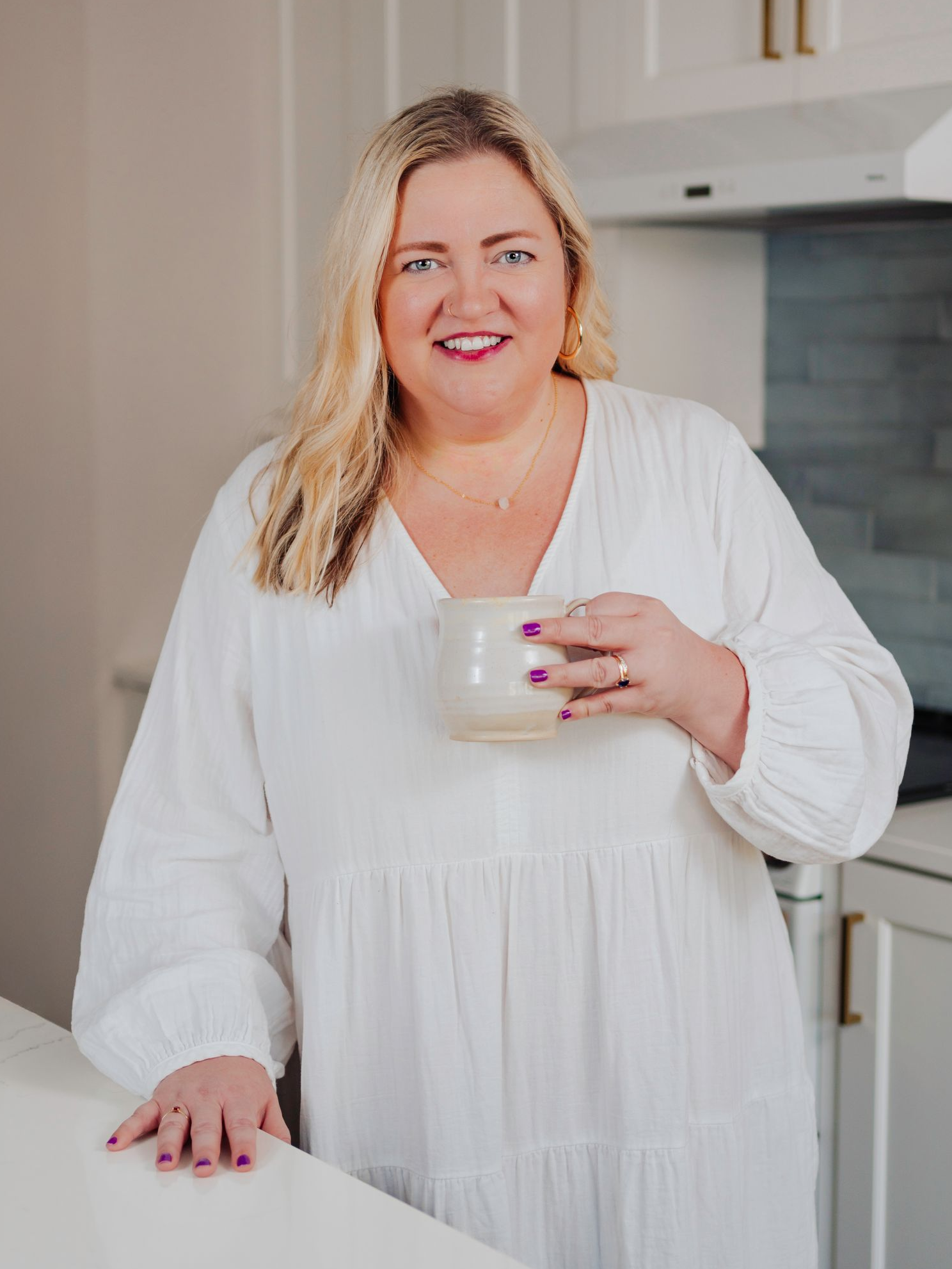 Erin Leigh Patterson smiles at the camera from her kitchen while drinking a cup of coffee