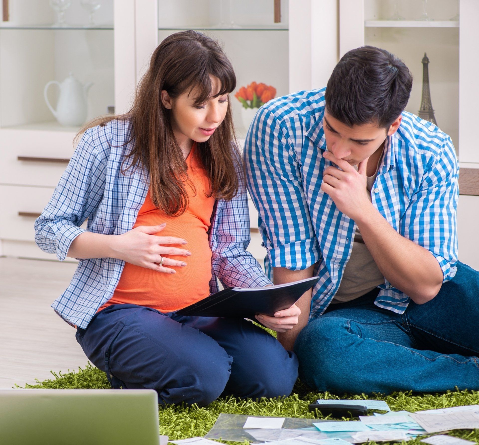 A couple sits together on a rug on the floor looking at bills and financial documents. The wife is pregnant and is feeling her belly with one hand and holding a folder in the other.