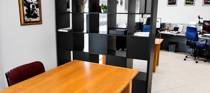 Interno di un ufficio con scaffali neri, documenti su un tavolo di legno e scrivanie con computer sullo sfondo.