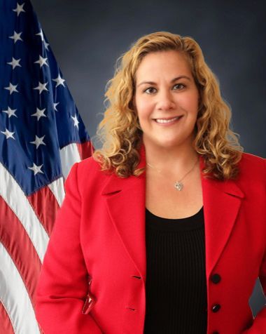 Woman in red blazer smiles in front of an American flag.