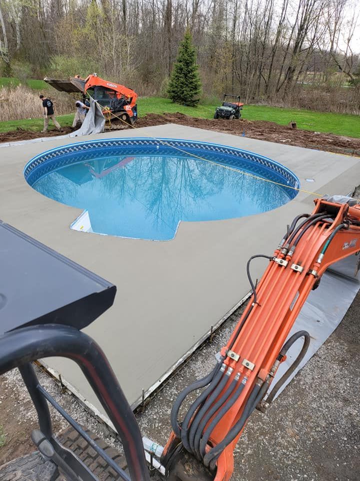 Concrete patio with pool, brown umbrella, picnic table, and fence in backyard.