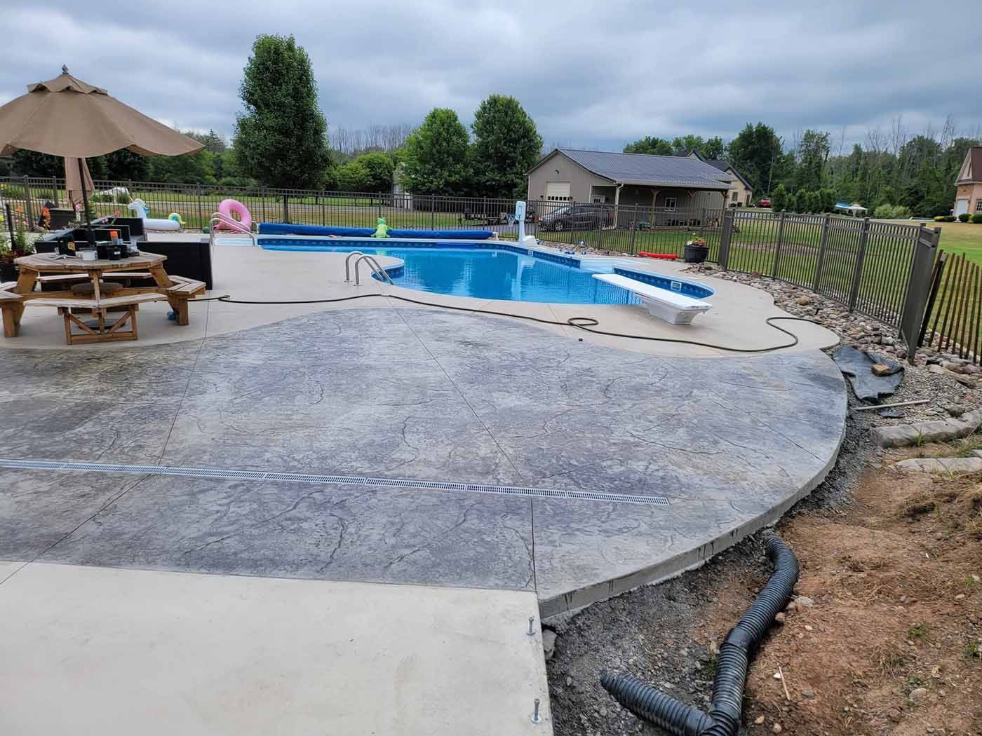 Concrete patio with pool, brown umbrella, picnic table, and fence in backyard.