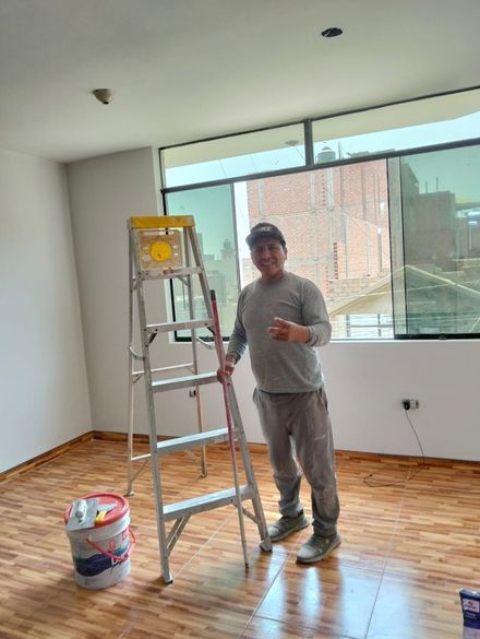 Una persona de pie sobre una escalera en una habitación vacía con suelo de madera, materiales de pintura y grandes ventanales.