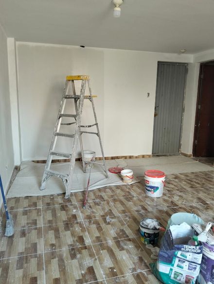 Habitación parcialmente reformada con escalera, materiales de pintura y paredes sin pintar sobre suelo de baldosas.