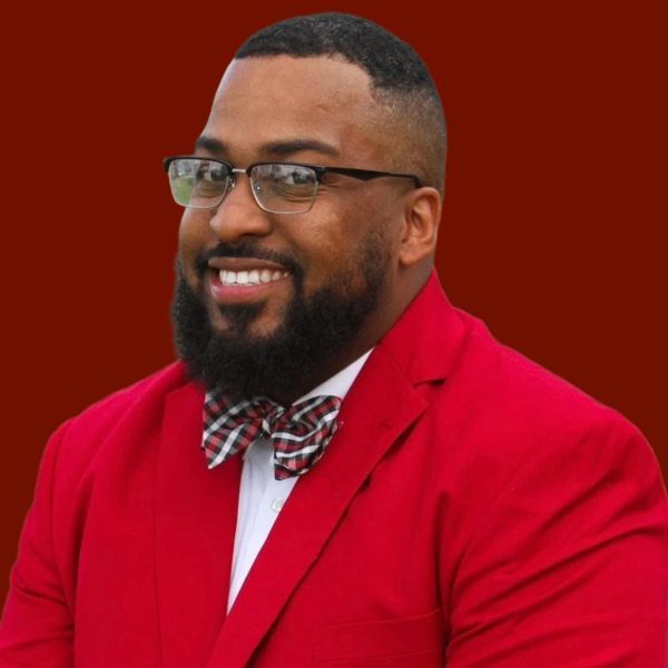 Smiling Black man in a red blazer and plaid bow tie, wearing glasses against a red background.