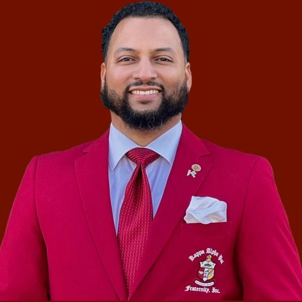 Man in red suit, red tie, smiling, with a fraternity logo.