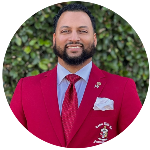 Man in red blazer, white pocket square, tie, and a pin, in front of a lake.