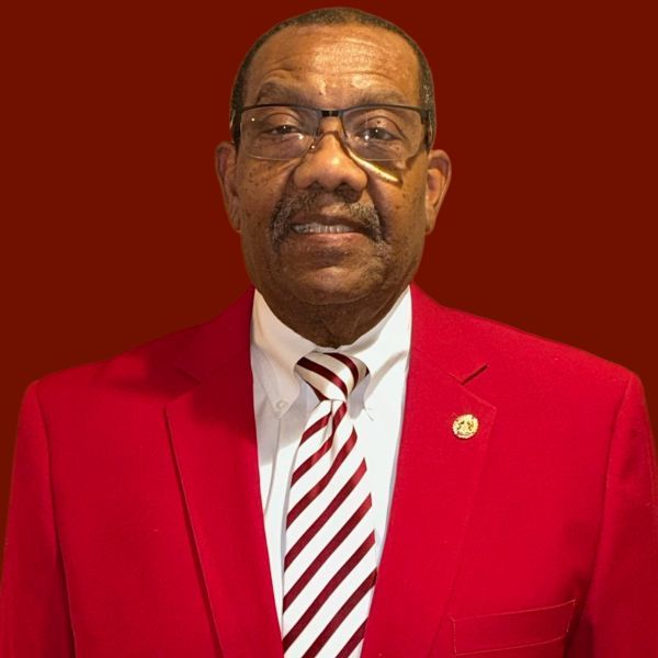 Man in red blazer, white shirt, and striped tie, wearing glasses, smiling, with a red background.