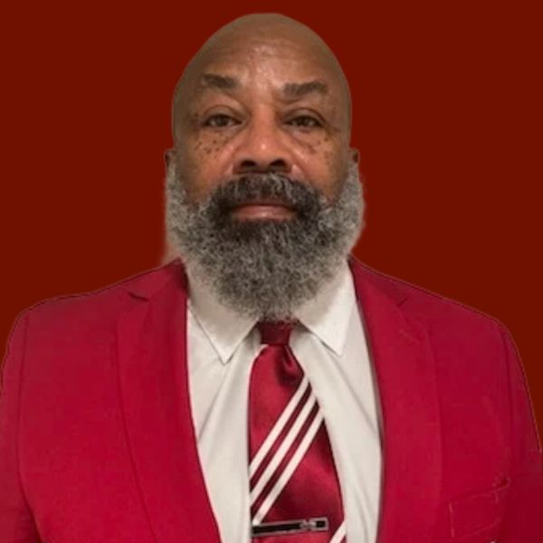 Man in a red suit, white shirt, and striped tie. He has a grey beard and is looking forward.