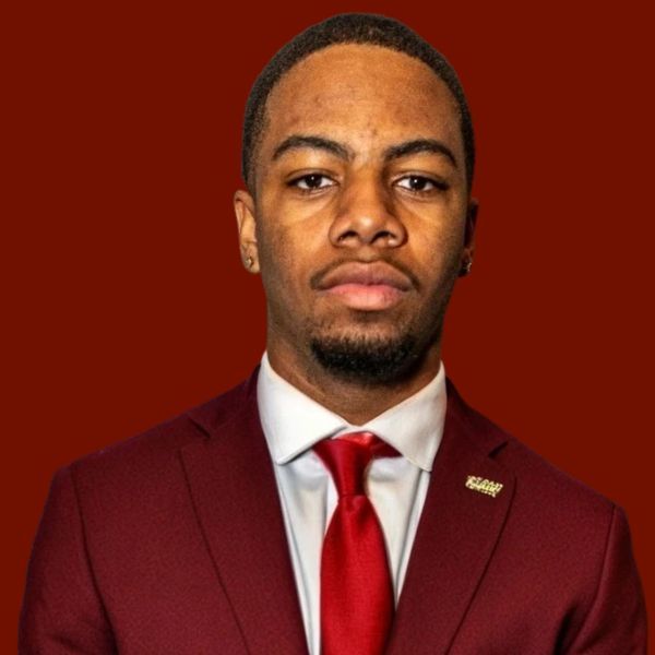 Man in maroon suit, red tie, and white shirt, against a maroon background.