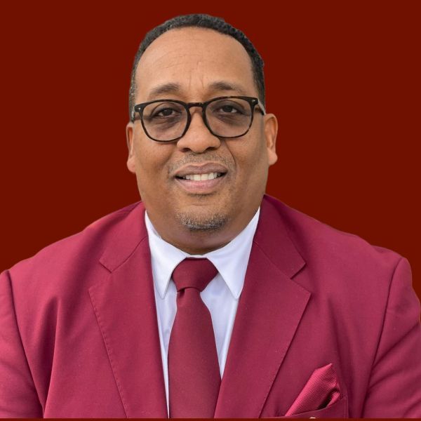 Man in maroon suit, glasses, and tie, smiling against a maroon background.