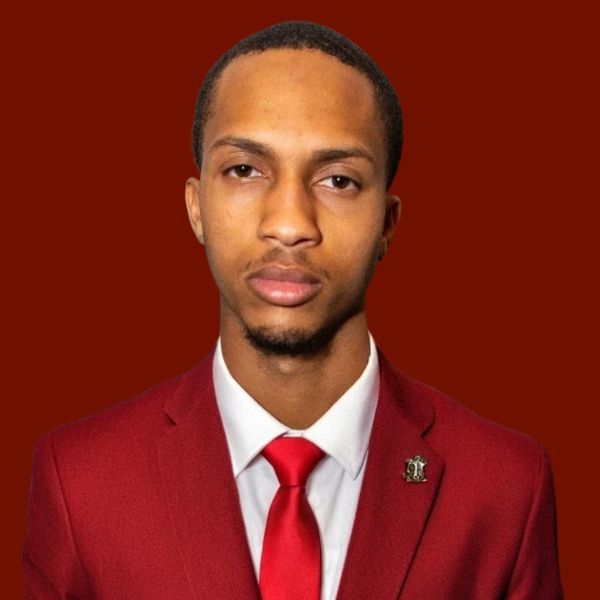 Man in red suit and tie against a red backdrop. He has a serious expression and a lapel pin.