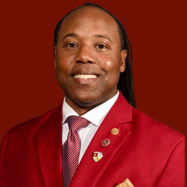 Man with dreadlocks in a red suit smiles at the camera, multiple pins on lapel.