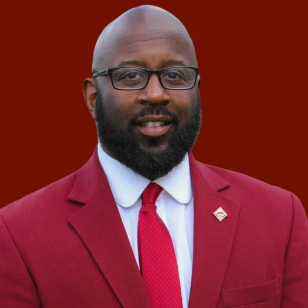 Man in red suit, glasses, and tie smiles against a red background.