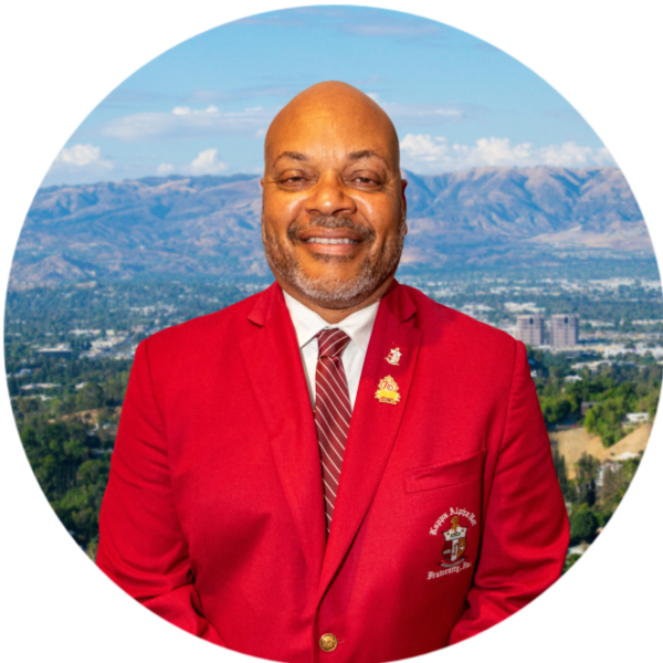 Bald man in a red blazer smiles, set against a background of green hills and mountains.