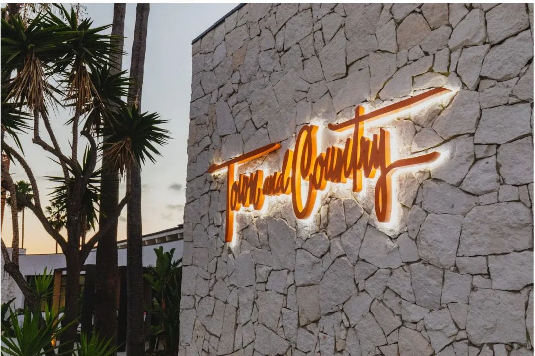 "Town and Country" sign on a stone wall, lit in orange against a neutral backdrop; palm trees in the background."