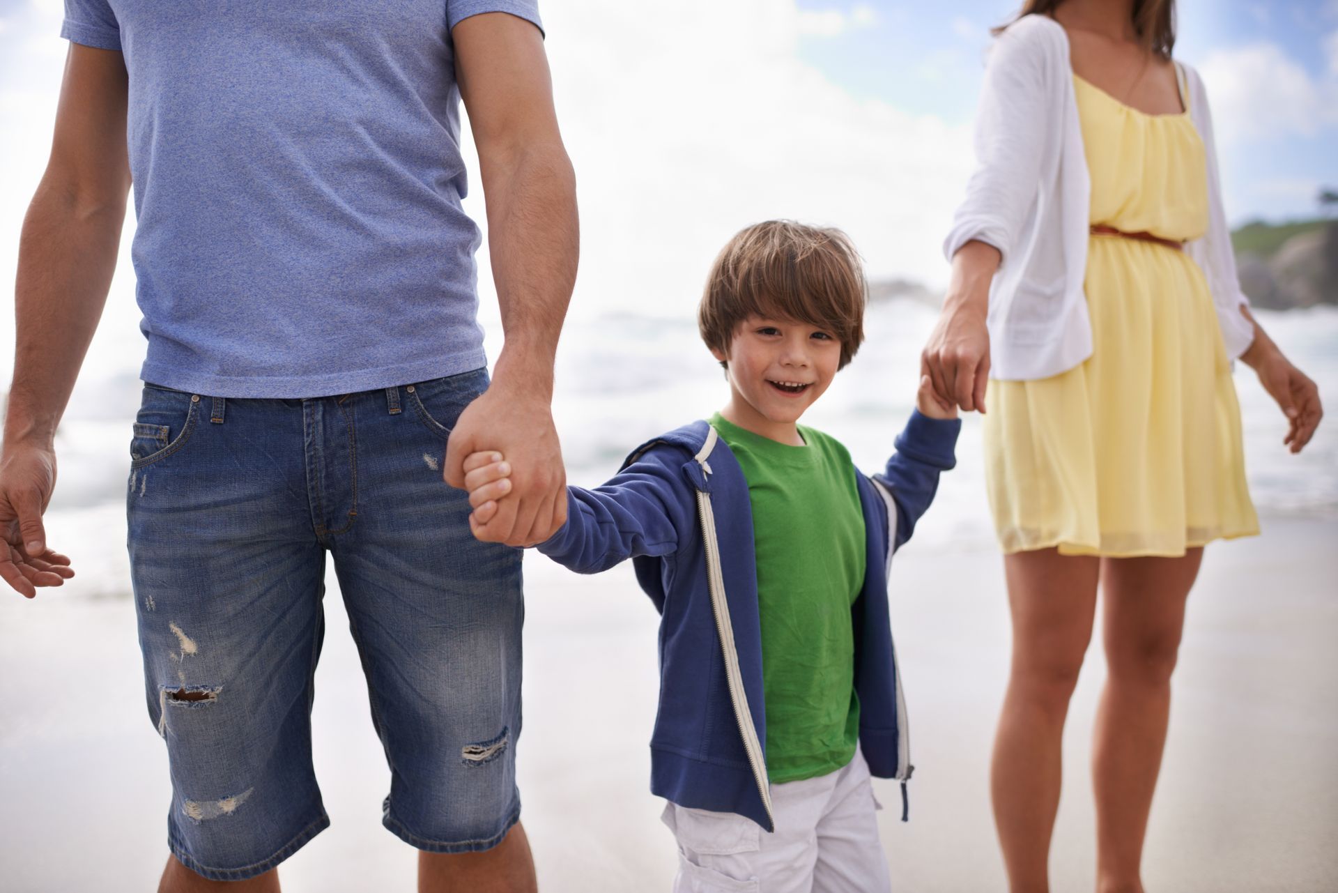 A child smiles while holding hands with two adults on a beach.