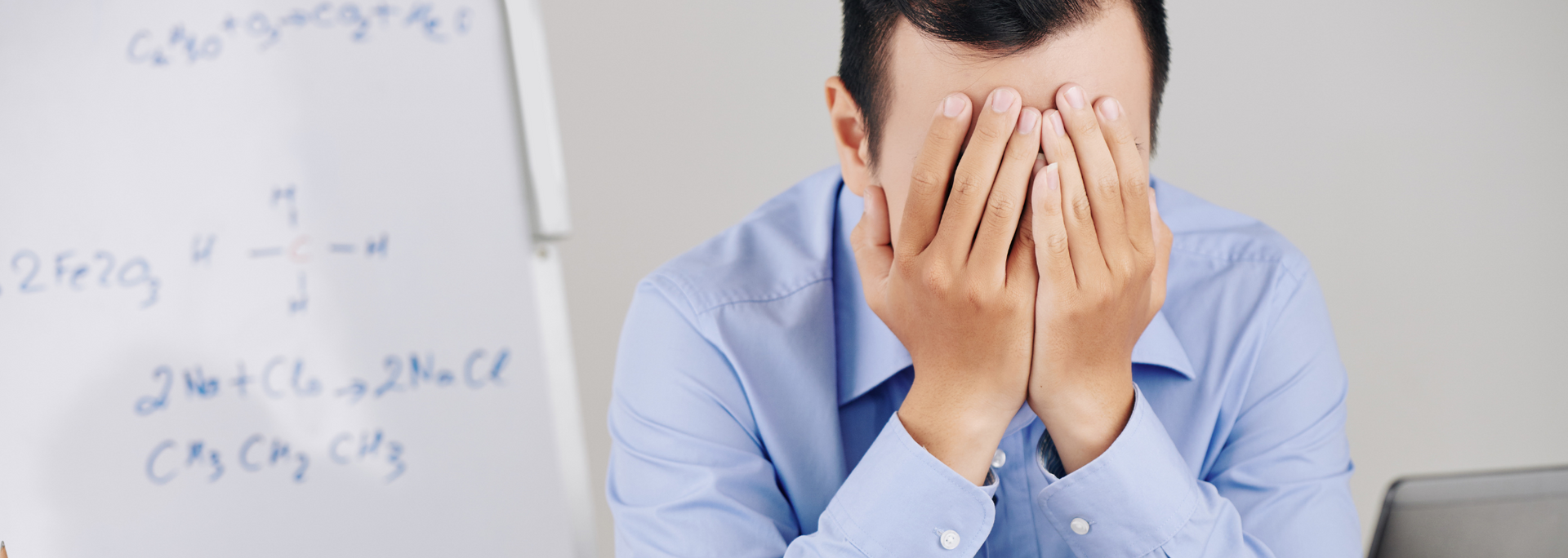 A man is covering his face with his hands in front of a whiteboard.