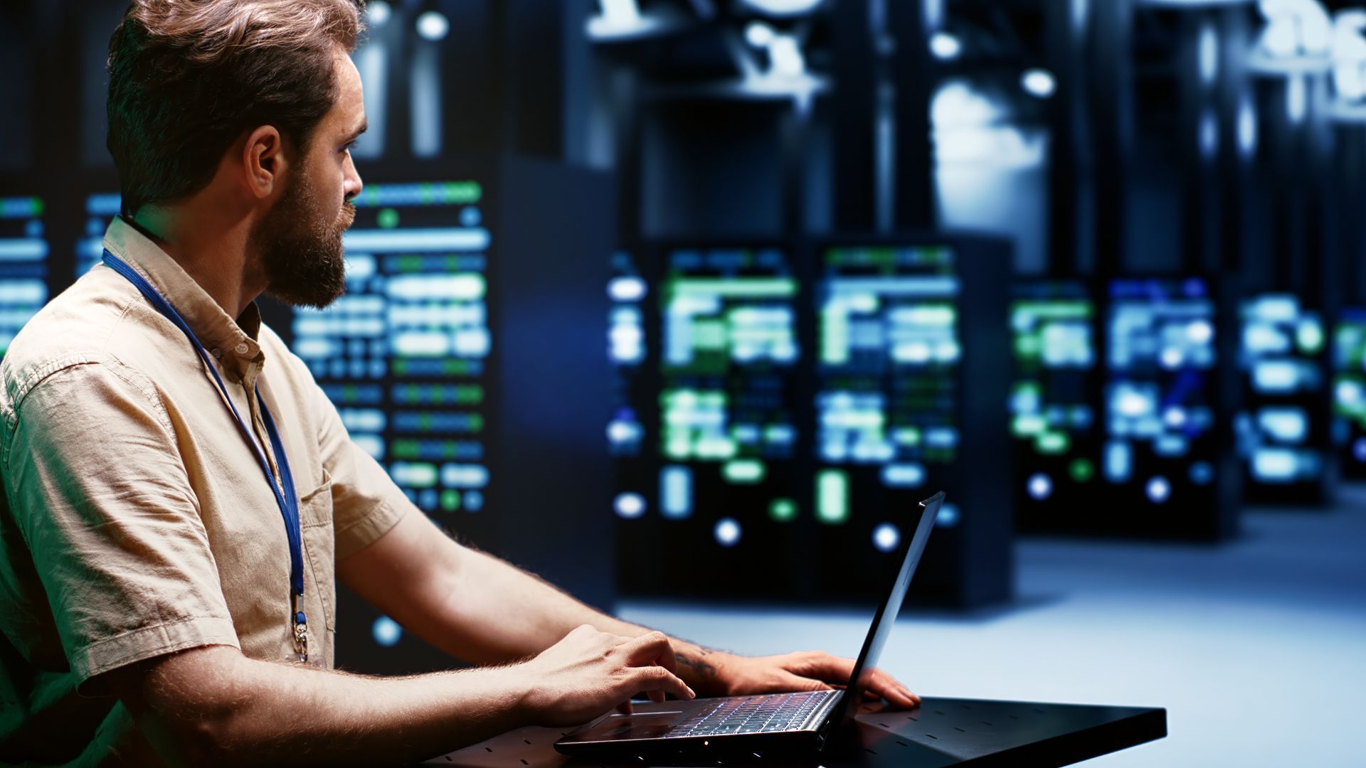 A man is sitting at a desk using a laptop computer in a server room.