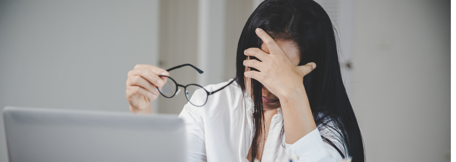 A woman is covering her face with her glasses while using a laptop computer.