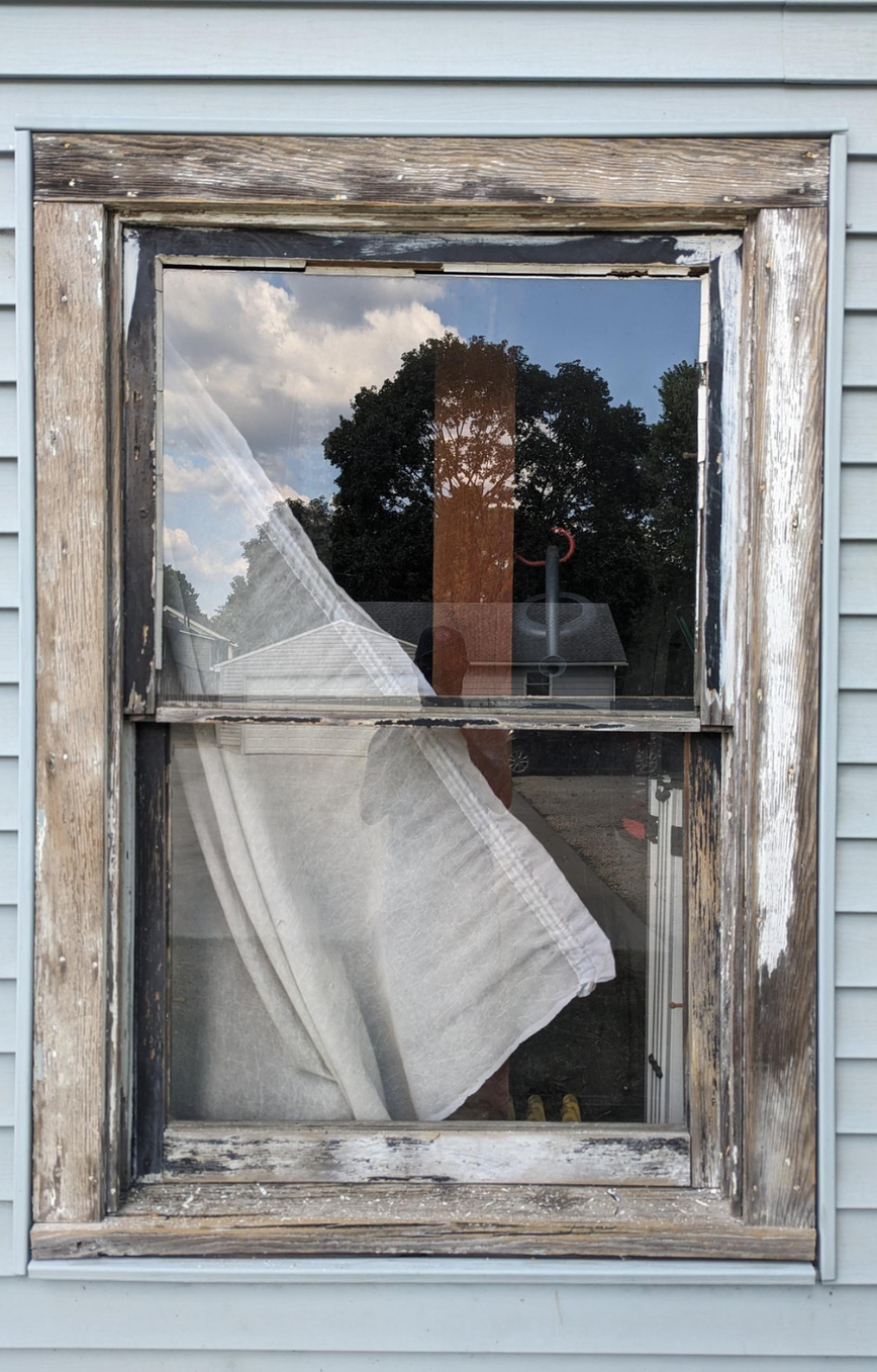 Weathered window with torn white curtains on a gray-sided house reflecting trees outside