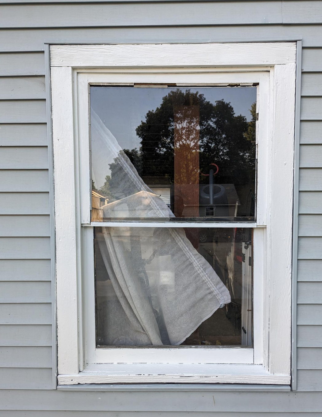 Damaged white-framed window with a broken lower pane and a hanging curtain on gray siding.