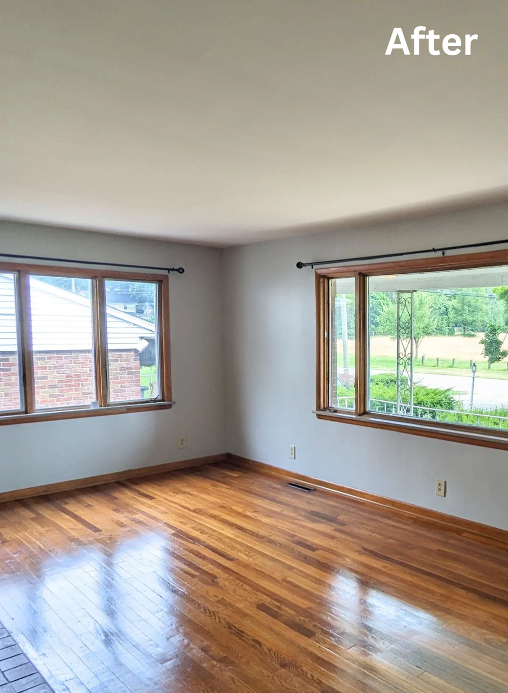 Empty living room with shiny hardwood floors, white walls, and large windows overlooking a green yard