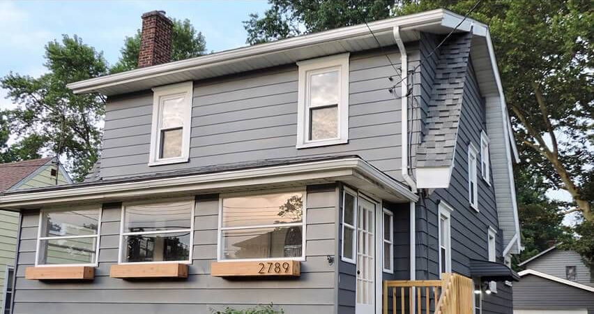 Gray two-story house with white trim, large front windows, and a small porch under a cloudy sky