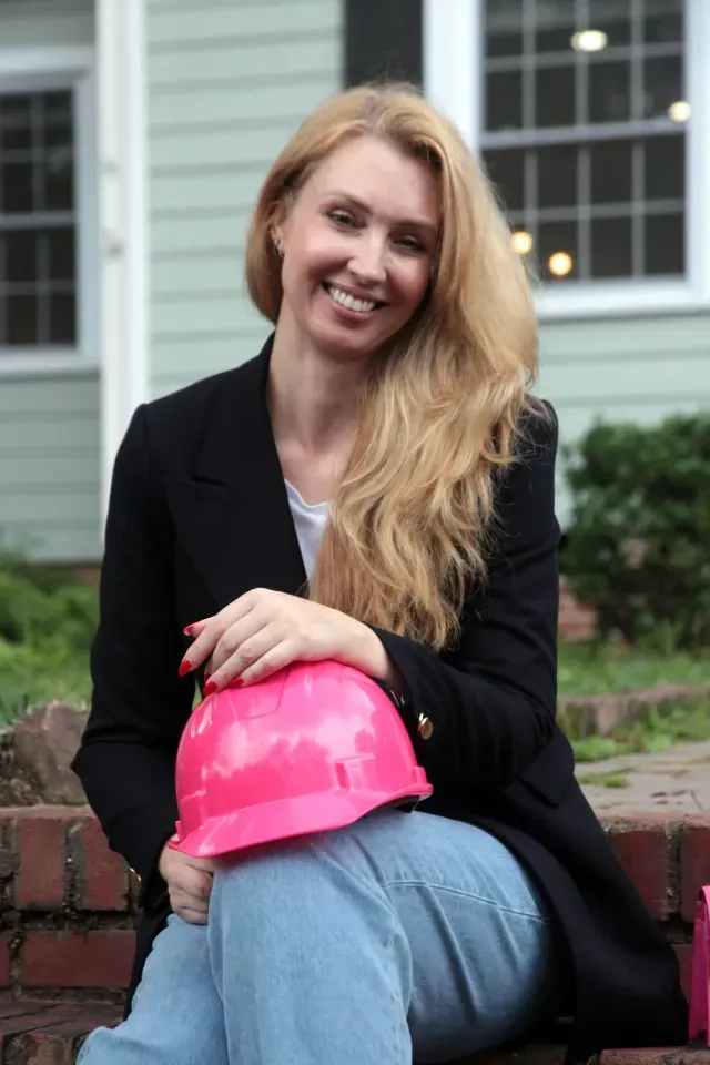 A woman is sitting on a brick wall holding a pink hard hat.