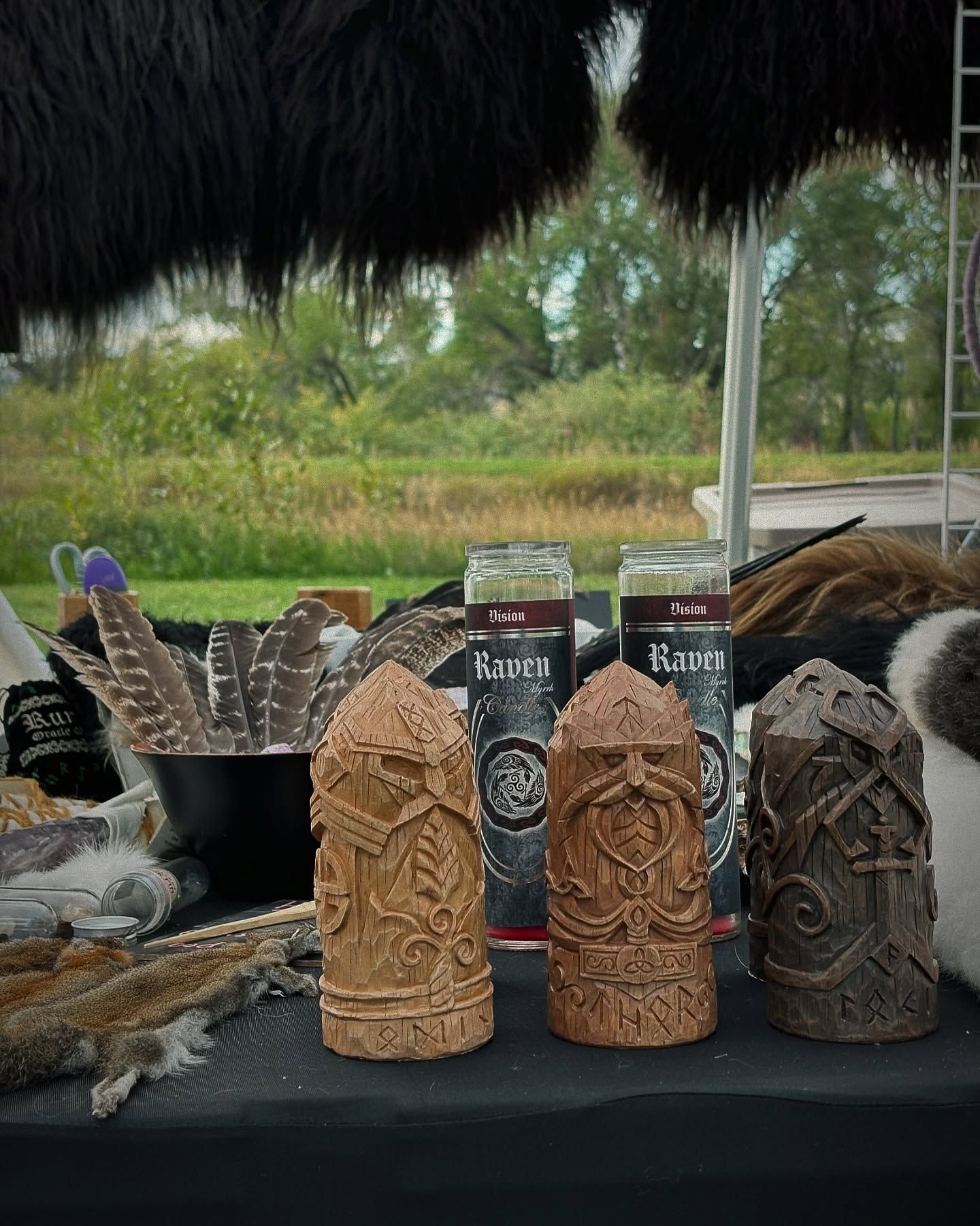 Table of Viking-themed goods: carved statues, candles, feathers, and furs, with a grassy outdoor background.