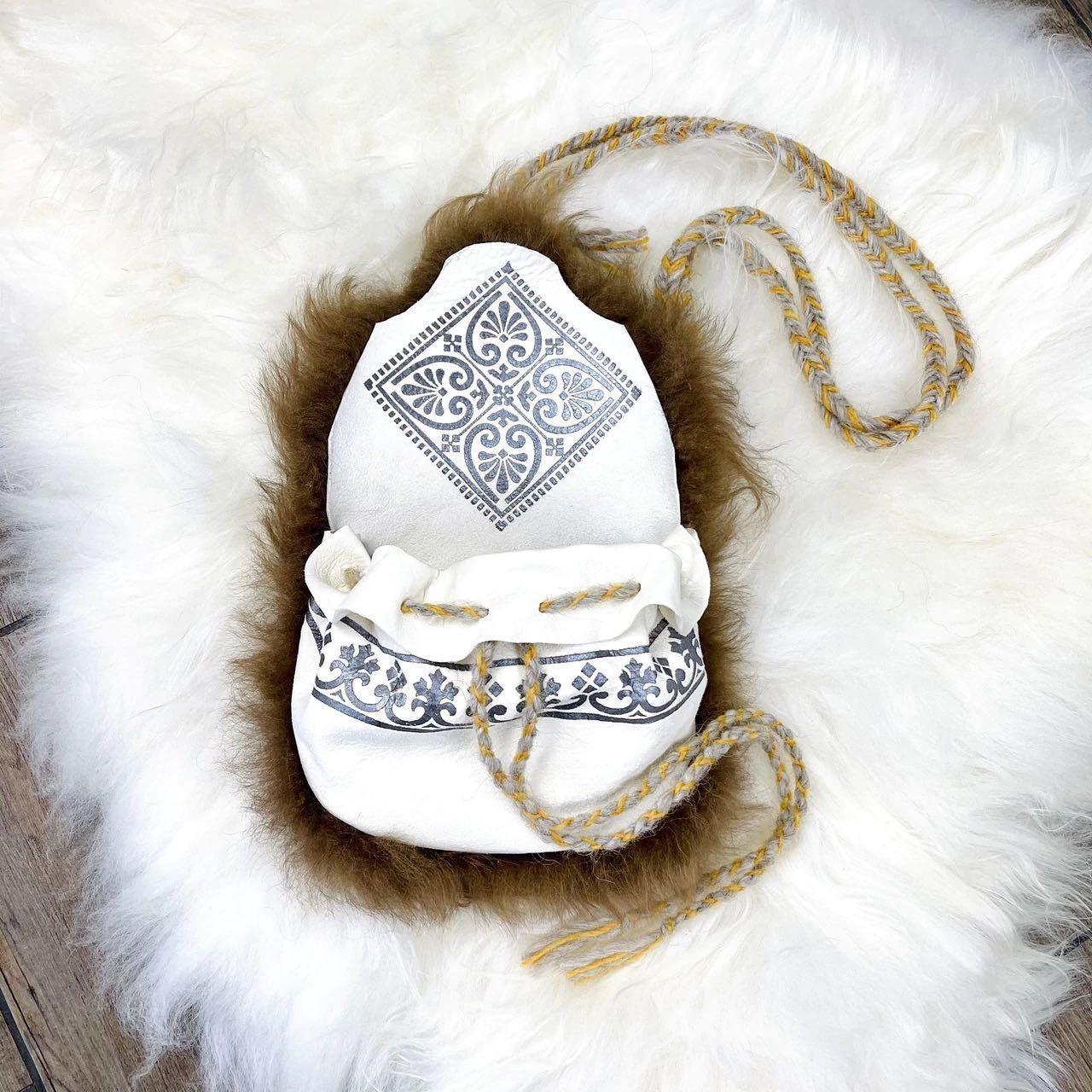 White pouch with black embroidered designs, drawstring, and strap, on a shaggy rug.