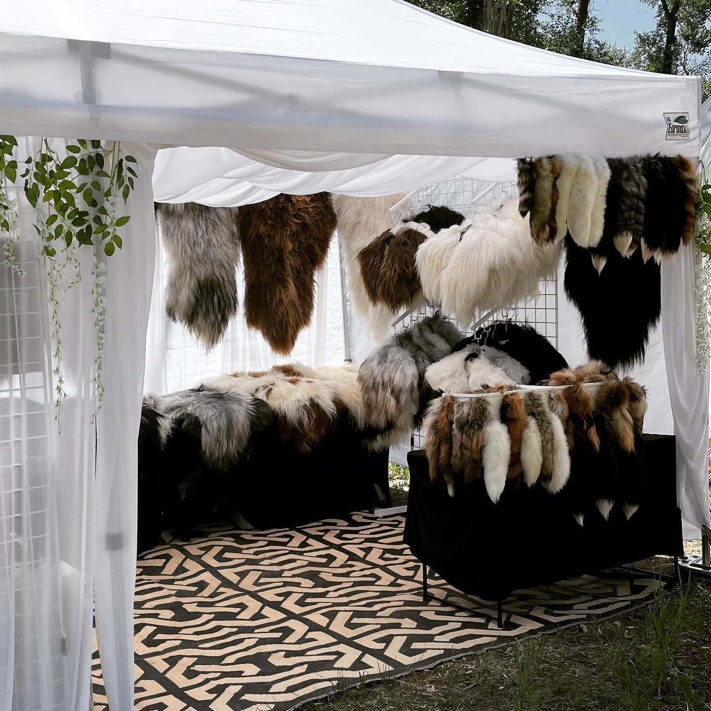 Fur rugs and pelts displayed in a white tent at an outdoor market, patterned rug on ground.
