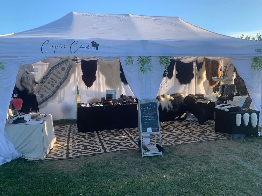 White tent with a vendor's booth displaying rugs, jewelry, and accessories; outdoor setting.