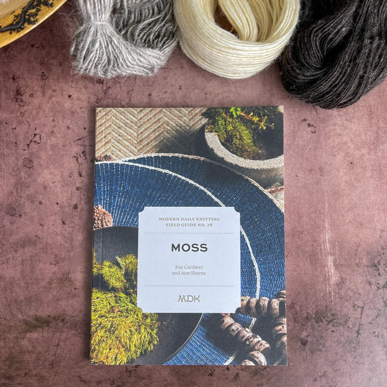 A book titled "Moss" with yarn samples on a rustic surface.