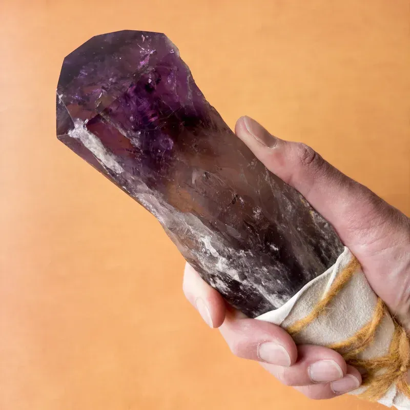 Hand holding a large, purple amethyst crystal wrapped in white fabric against an orange background.