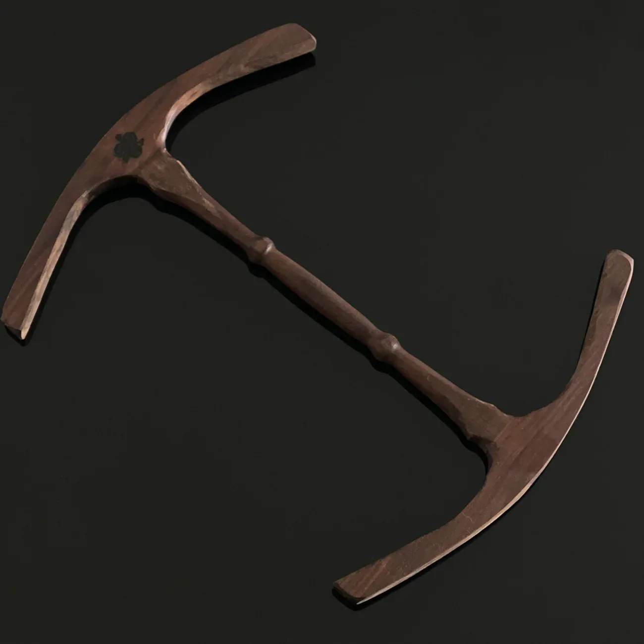 Wooden double-pronged weapon with central handle, possibly a pickaxe. Dark background.