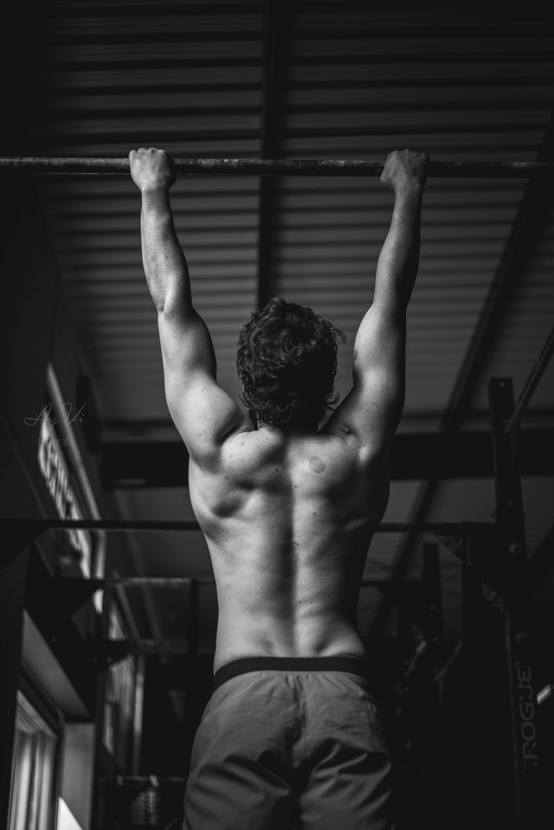 Person doing a pull-up, viewed from behind in a gym, with arms extended overhead.