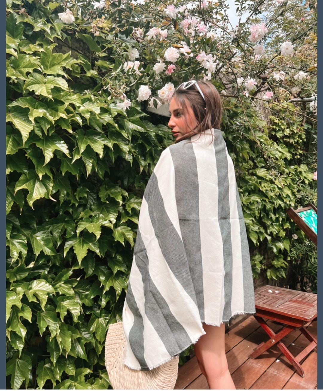 Mujer envuelta en una toalla de rayas grises y blancas, de pie cerca de vegetación y flores.