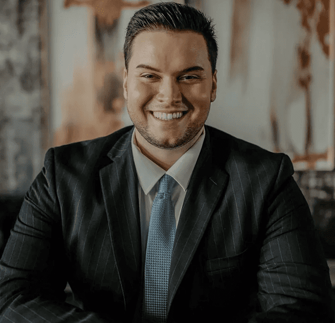 Man with dark skin, wearing a blue suit and red tie, smiling. White background.